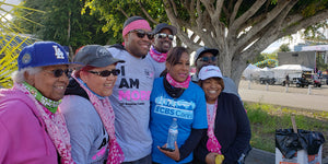 Group of people wearing pink scarves and hats, standing outdoors with trees and a road in the background during community cancer walk event.